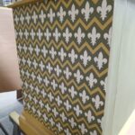 Wooden cabinet front covered with dark green wallpaper featuring white fleur-de-lis and gold chevron stripes.