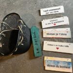 Black wire basket on a rough concrete floor beside a teal vertical sign reading 'Le jardin', and several hanging wooden plaques with French messages about moms, including 'Maman je t'aime', 'Maman tu es la plus belle', and 'Maison du bonheur' (some plaques feature small red hearts).