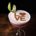 Pink cocktail in a coupe glass with a foamy top, cinnamon lettering spells 'LOVE' and a mint leaf garnish on the rim