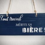 Blue wooden hanging sign with white chalk text reading 'Tout travail mérite sa bière!' mounted on a white wall with rope handles.